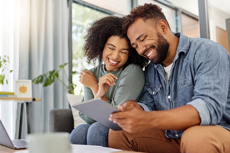 A smiling couple sitting on the couch looking at a tablet